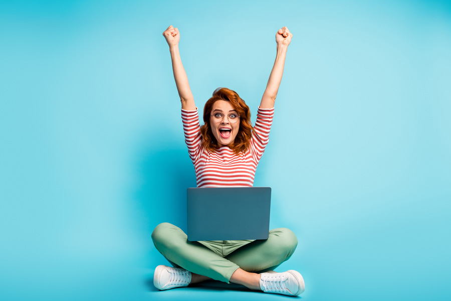 young woman celebrating with hands in the air sat cross legged with laptop open on her lap