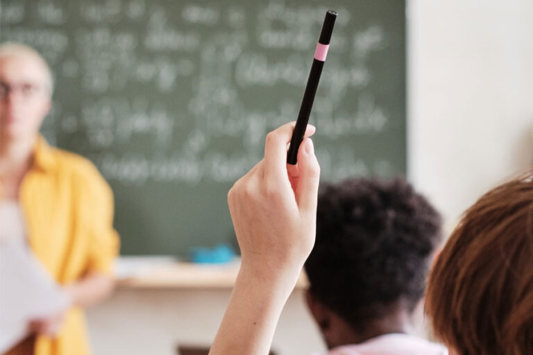 close up of someones hand up in a classroom with teacher is background